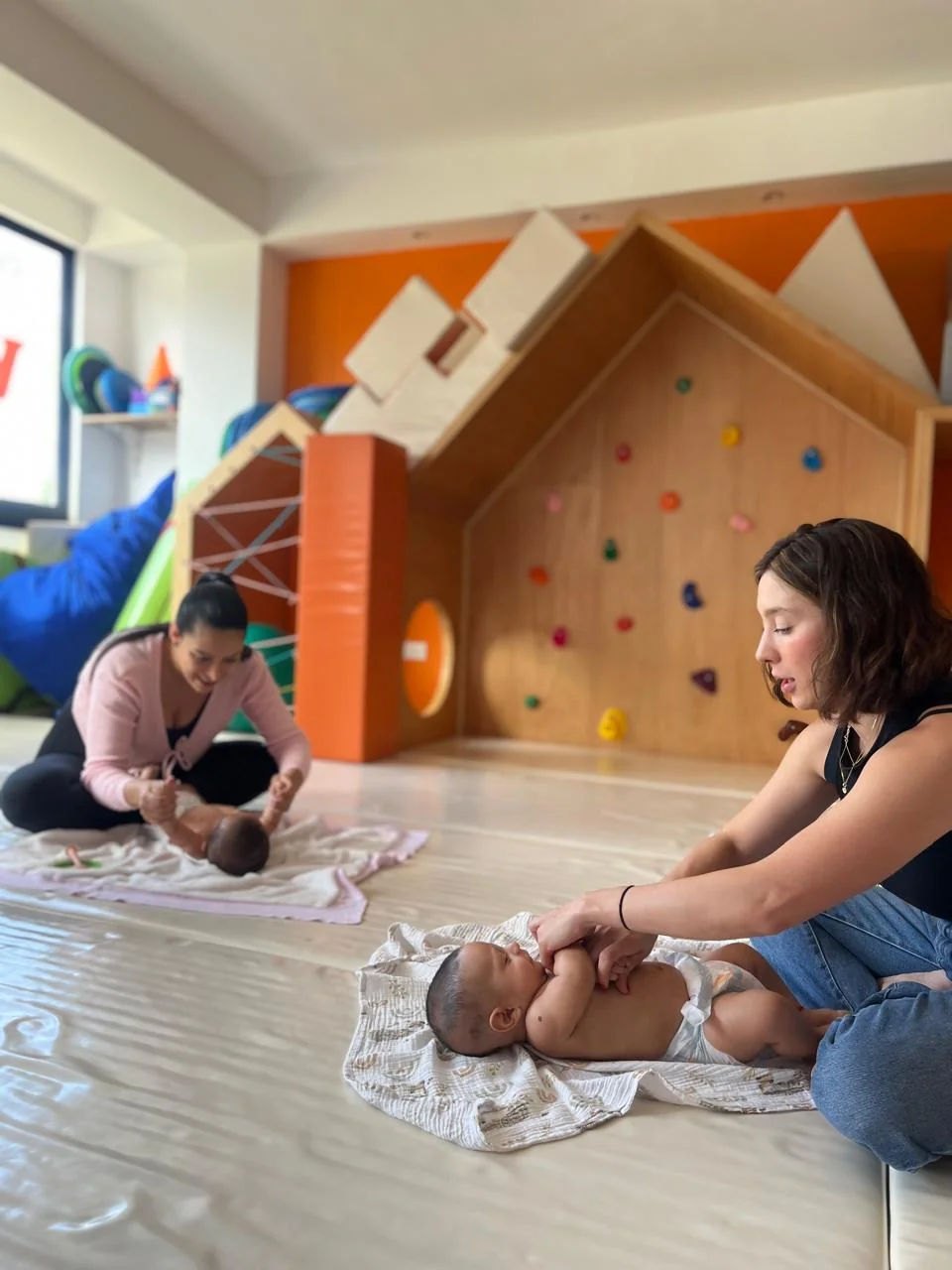 Dos mamás dando masaje a sus bebés sobre mantas, frente a la casita del aula